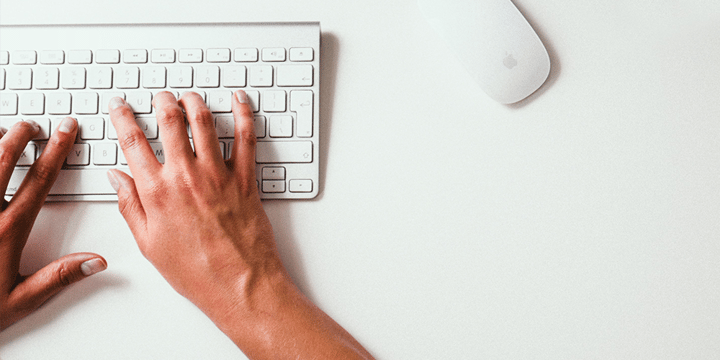 Half of a white computer keyboard is shown with a hand typing on it. There is a white computer mouse on the right of the image.