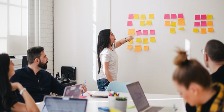 Asian woman presents in a meeting room. She is pointing towards post-it notes. There are 4 people sat at the table with laptops watching her.