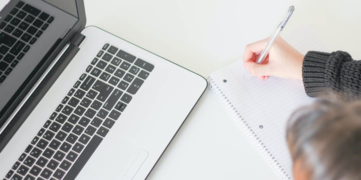 Half of a laptop keyboard is shown on the left hand side of the image. On the right is a hand holding a pen writing on a notepad. 