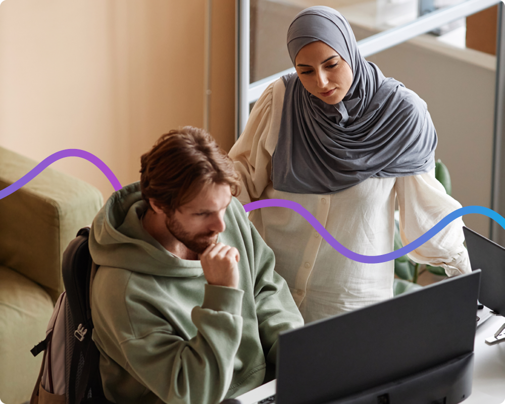 Image of a man sat at a desk looking at his laptop and his colleague is standing next to him.