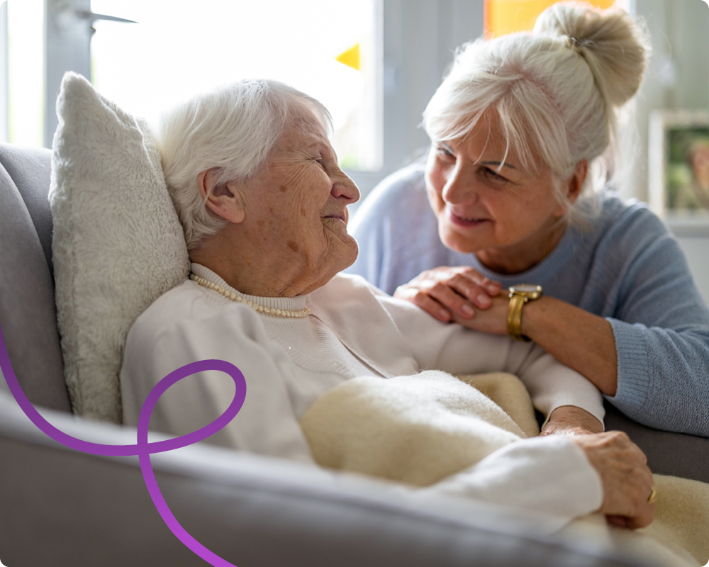 Image of an old woman, sat in her chair smiling, whilst her daughter is crouched down next to her