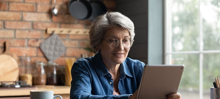 Marge, age 64, smiles whilst sitting at a table holding her digital tablet.