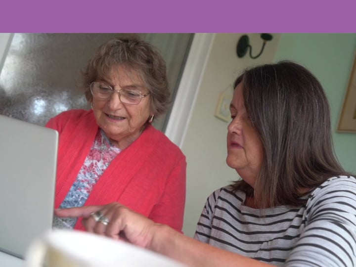 An elderly woman on the left in a red blouse sat next to a middle aged woman in stripey black and white top and both are looking at a laptop computer.