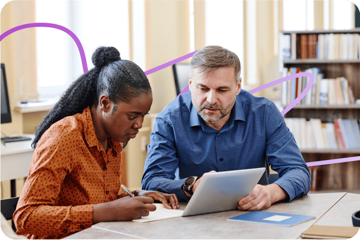 A caucasian man teaching a Black woman how to use a tablet device. The woman is listening and taking notes.