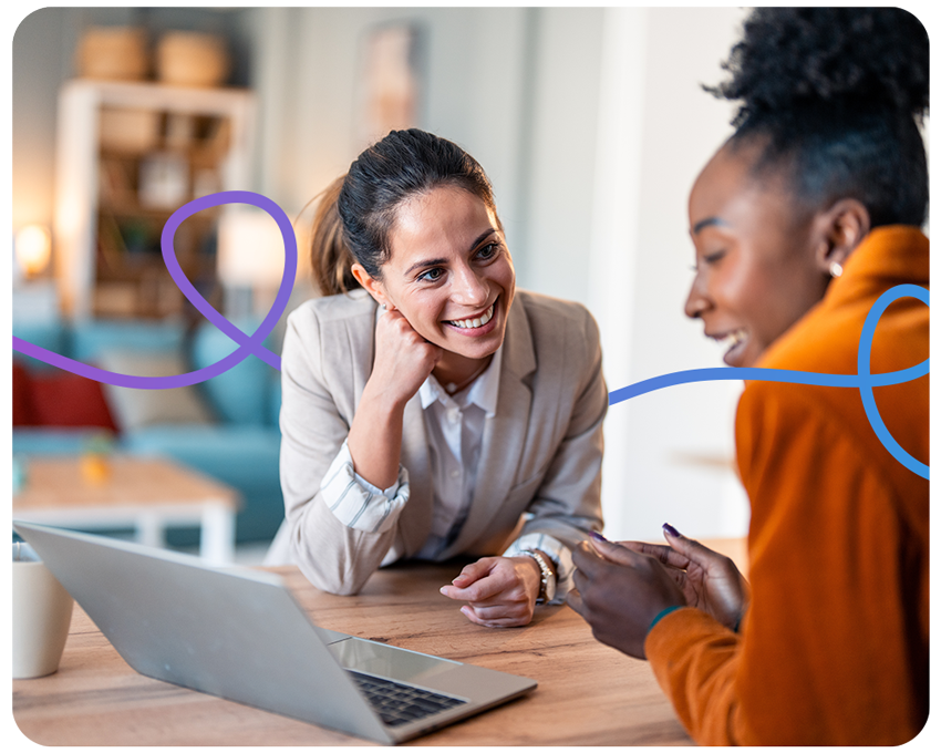 Two professional women, one in beige blazer and another in orange turtleneck, engaged in animated discussion at wooden table with laptop in sunlit home office