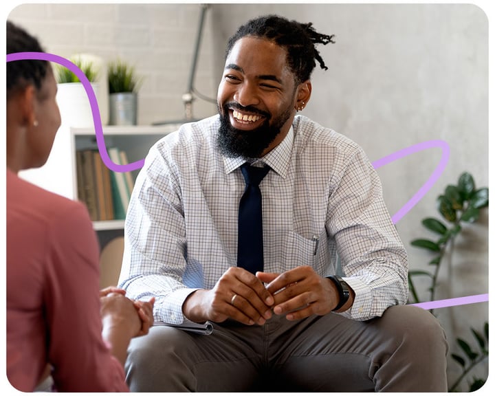 Two people sitting on a cound talking. The person on the left is cut out of the shot but we see the person on the right looking at them and smiling.
